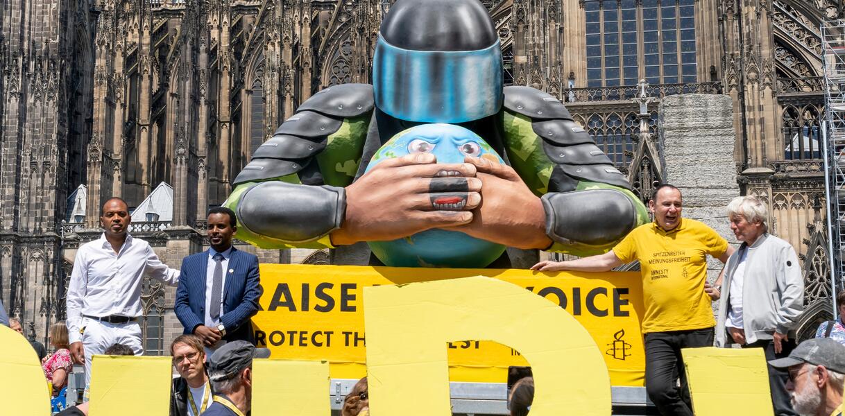 Das Foto zeigt eine Gruppe Menschen, die vor und neben einer großen Figur aus Pappemaché steht: Eine Person mit Schutzhelm und Schutzausrüstung drückt einer stilisierten Erdkugel beide Hände auf den Mund, um sie am Sprechen zu hindern. 