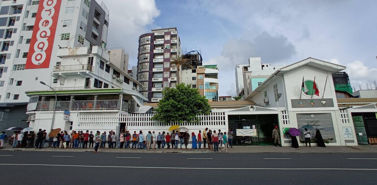 Schlange aus wartenden Menschen am Straßenrand vor einem Gebäude. Im Hintergrund befinden sich weitere Gebäude.
