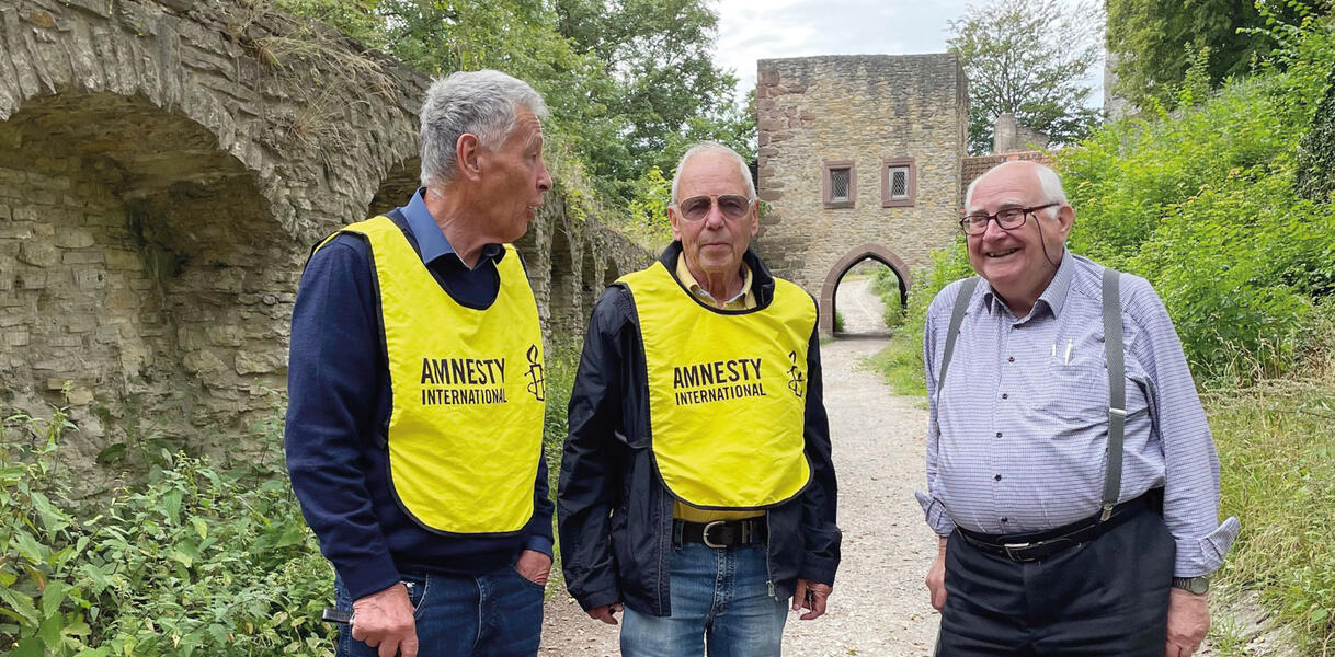 Drei Männer stehen auf einem Trampelpfad, rechts von ihnen eine altertümliche Mauer, hinter ihnen ein Torbogen. Zwei Männer tragen Schutzwesten mit der Aufschrift "Amnesty", ein älterer Mann ganz rechts hält eine Gehstütze und lächelt.