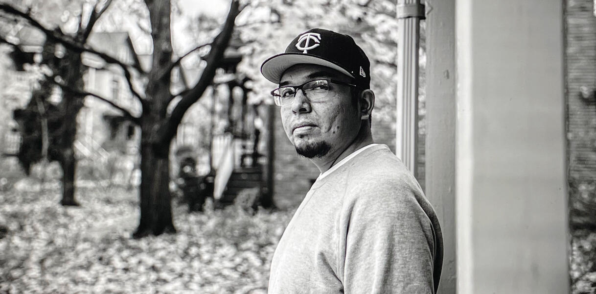 Ein Mann mit Sweat-T-Shirt und Baseball-Kappe trägt eine Brille und steht in einem Hauseingang, im Hintergrund stehen Bäume.