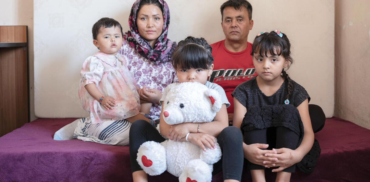 Eine afghanische Familie sitzt auf einer Matratze mit Frotteebezug, die auf dem Boden liegt in einem Zimmer, an die Eand hinter ihnen ist eine weitere Matratze gelehnt, die Mutter mit Kopftuch hält ein Kleinkind halb auf dem Schoß, neben ihr sitzt der Vater, vor ihnen ihre zwei Töchter auf der Kante der Matratze, eine versteckt ihr Gesicht halb hinter einem großen Teddybären, die andere hat ihre Beine angezogen und ihre Arme darum geschlungen.