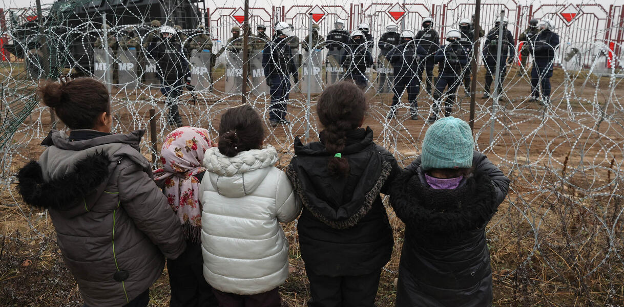 Fünf Kinder in Winterkleidung stehen mit dem Rücken zur Kamera vor einem Stacheldrahtzaun. Auf der anderen Seite des Zaunes stehen Sicherheitskräfte mit Helmen und Schutzschilden.