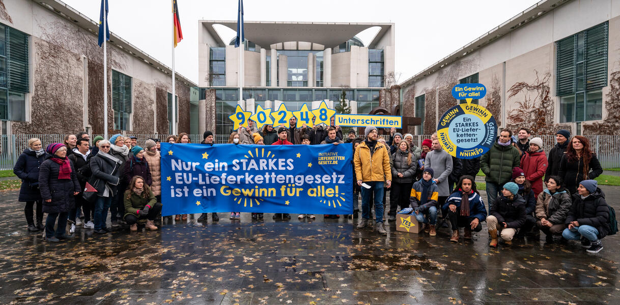 Eine Gruppe von circa 50 Personen steht vor dem Kanzleramt. Einige halten ein großes Banner vor sich. Auf dem Banner steht: "Nur ein starkes EU-Lieferkettengesetz ist ein Gewinn für alle".