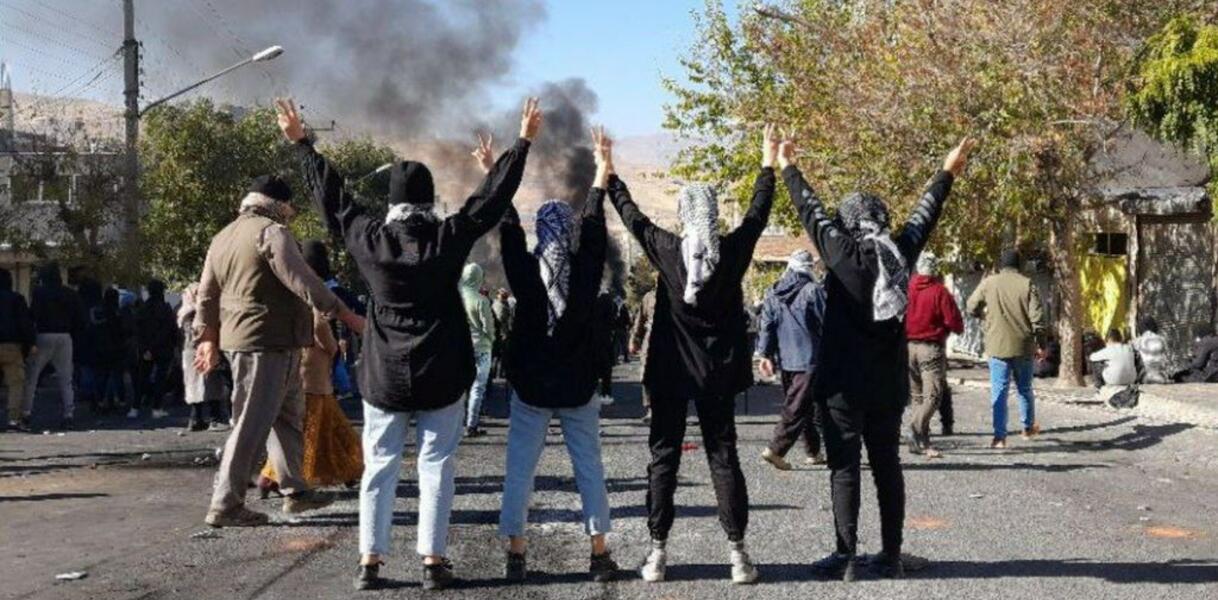 Vier Frauen mit Kopftüchern stehen nebeneinander auf einer Straße, Rauch steigt auf, sie recken ihre Arme in die Luft, es ist ein Straßenprotest.
