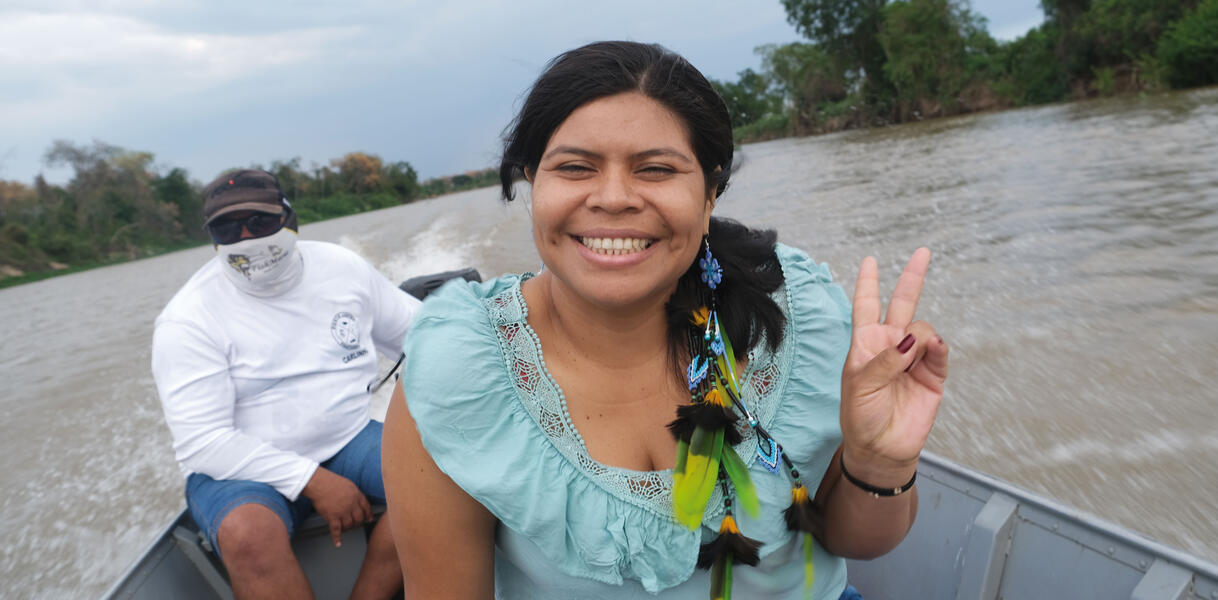 Eine indigene Frau sitzt auf einem Motorboot, lächelt, macht mit ihrer linken Hand das "Victory"-Zeichen, hinter ihr sitzt ein Mann, der das Boot steuert, im Hintergrund der Fluss und Bäume am Ufer.