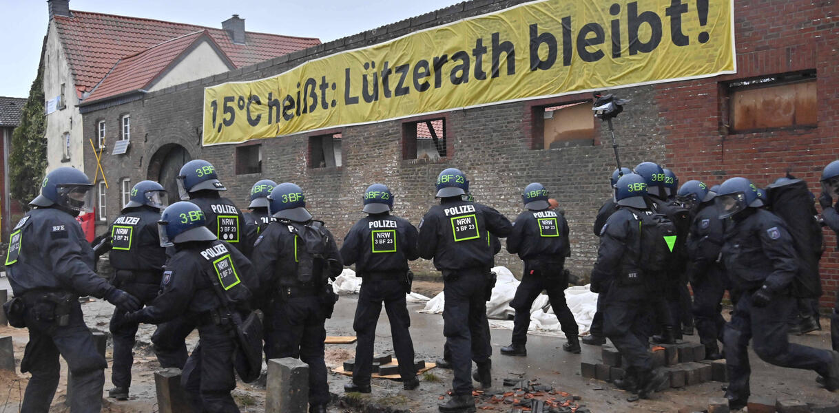 Das Bild zeigt im Vordergrund Polizisten, im Hintergrund ein Banner an einer Hauswand "Lützerath bleibt"