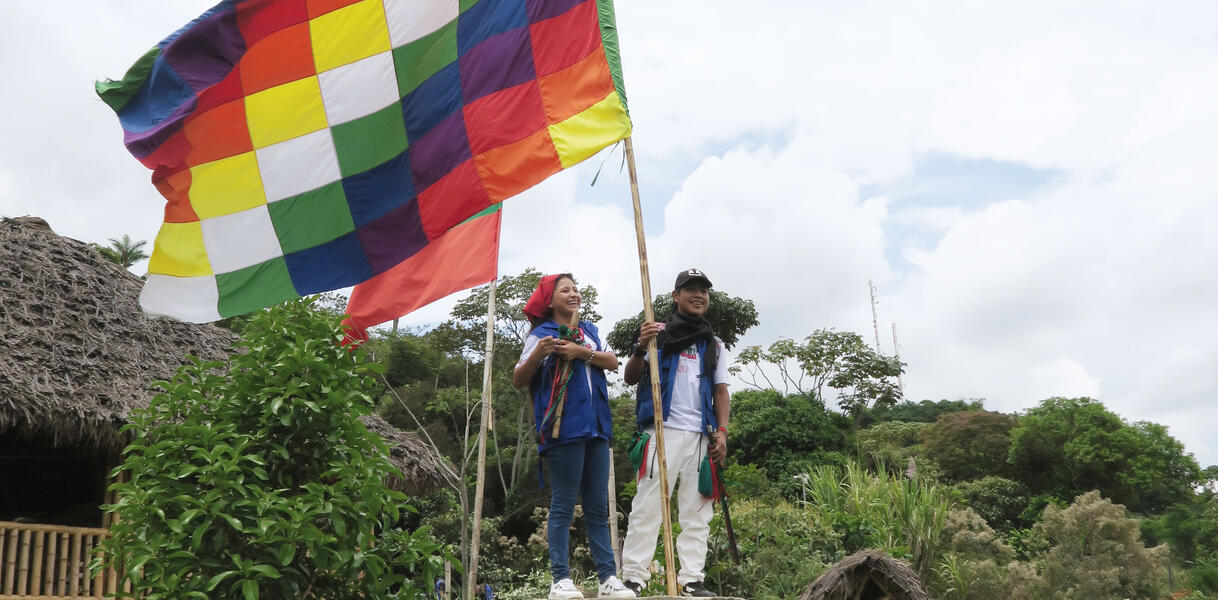 Indigene stehen vor einer Hütte an einem Waldstück und hissen eine Flagge, die im Wind weht; sie lächeln dabei.