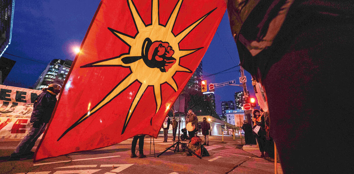 Menschen stehen auf einer Straße, jemand trommelt vor einem Lautsprecher, sie demonstrieren, eine Flagge mit einer Sonne und in deren Mitte dem Profil eines indigenen Mannes wird von jemandem gehalten.