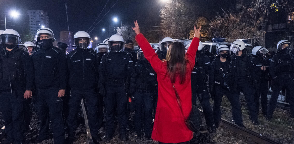 Das Bild zeigt eine Frau, die vor mehreren Polizisten steht und beide Hände in die Höhe hält