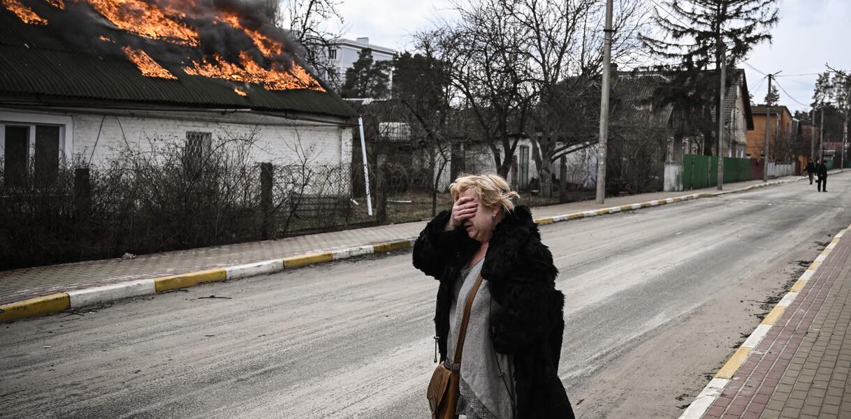 Eine Frau vor einem brennenden Haus hält sich entsetzt die rechte Hand vor ihr Gesicht.