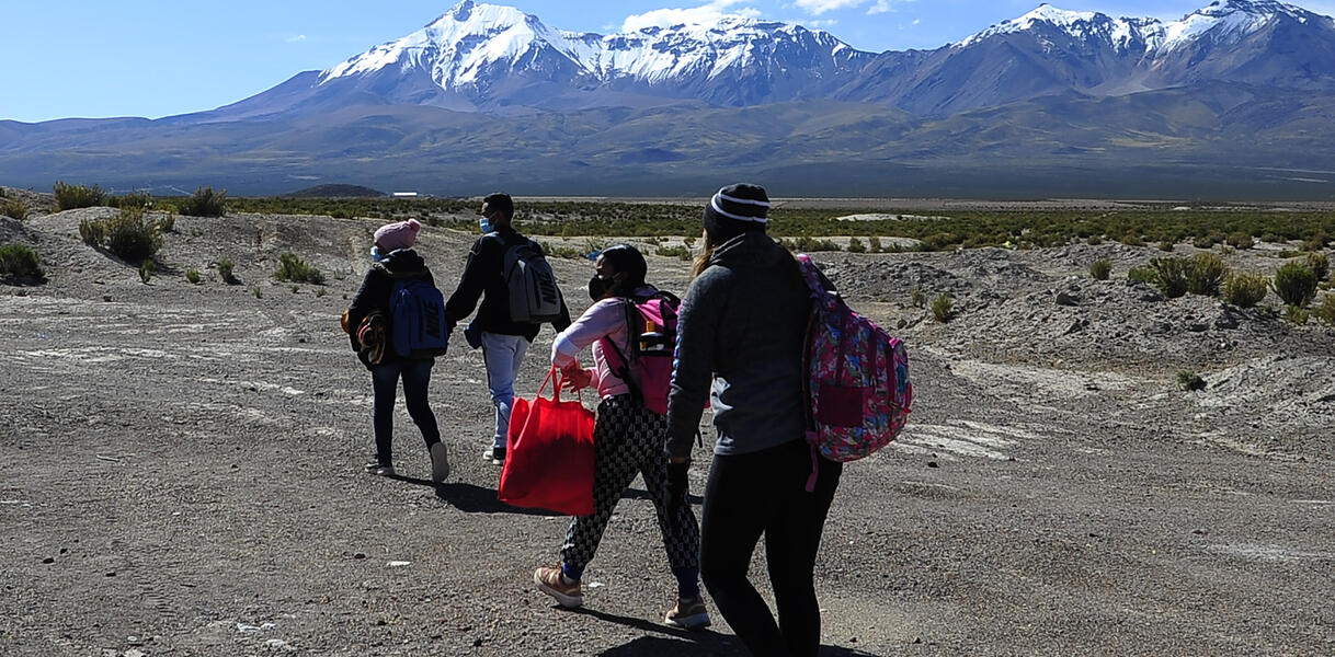 Das Bild zeigt mehrere Personen, die mit Taschen durch eine karge Landschaft gehen, im Hintergrund sind hohe Berge zu sehen.