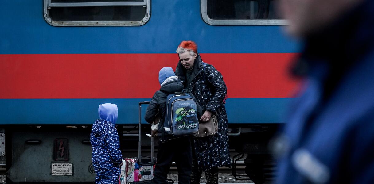 Das Bild zeigt einen Zug im Hintergrund, davor Kinder und Jugendliche mit Taschen
