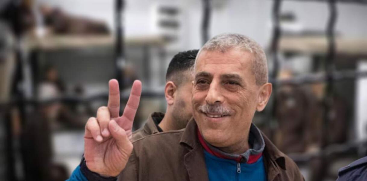 Ein älterer Mann mit kurzen grauen Haaren lächelt und formt mit Zeige- und Mittelfinger ein "V", es ist Walid Daqqah. 