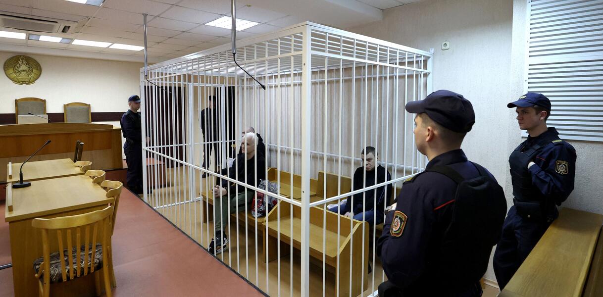 Drei Männer sitzen in einem Käfig in einem Gerichtssaal, um den Käfig herum stehen uniformierte Sicherheitskräfte, vier Männer.