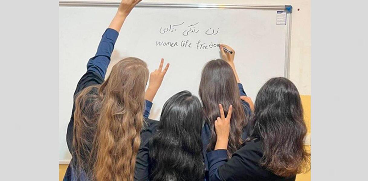 Das Foto zeigt vier Mädchen bzw. junge Frauen mit langen Haaren von hinten fotografiert. Drei von ihnen heben eine Hände und formen mit Zeige- und Mittelfinger das Victory-Zeichen. Eine von ihnen schreibt auf die Tafel.