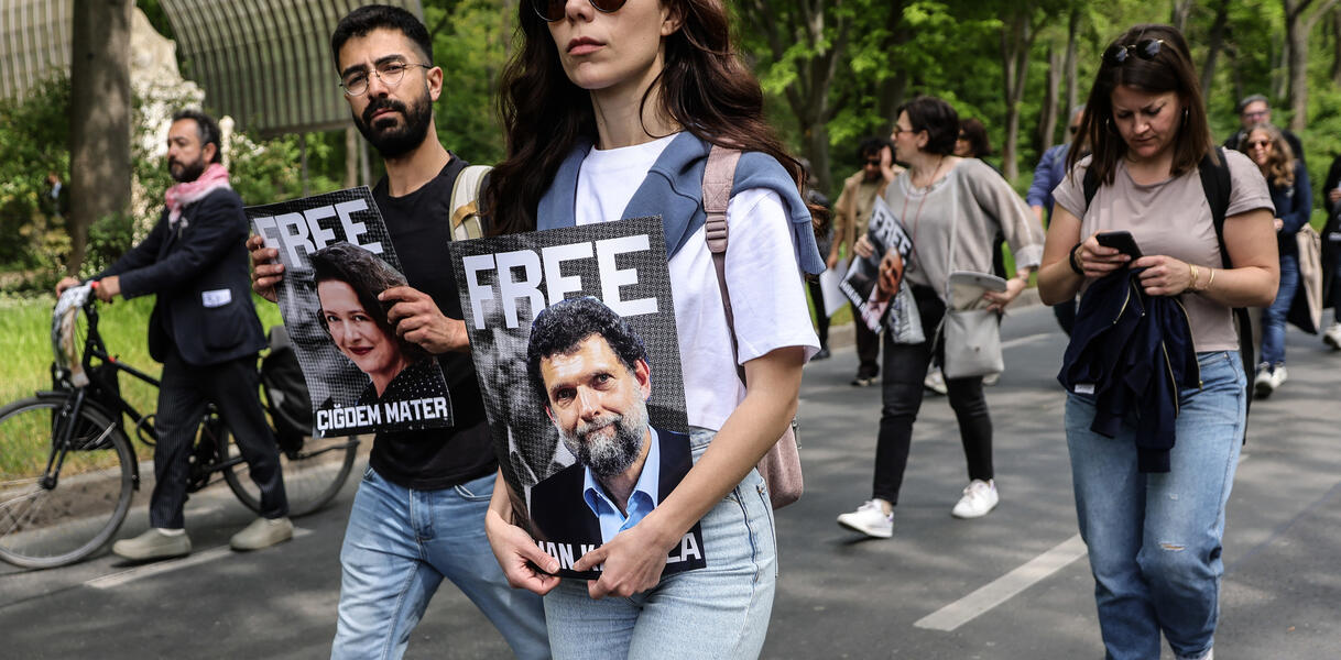Das Foto zeigt mehrere Personen auf einer Straße in einem Protestzug. Sie halten Porträtfotos vor sich. Eines der Fotos zeigt Osman Kavala.