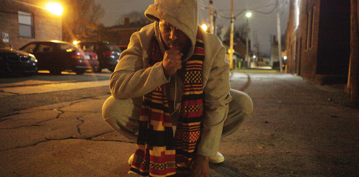 Ein Mann trägt eine Jogginghose, Sneaker, einen Hoodie und einen Schal, er hockt auf einer Straße bei Nacht, im Hintergrund Straßenlaternen und Autos, Gebäude aus Ziegeln.