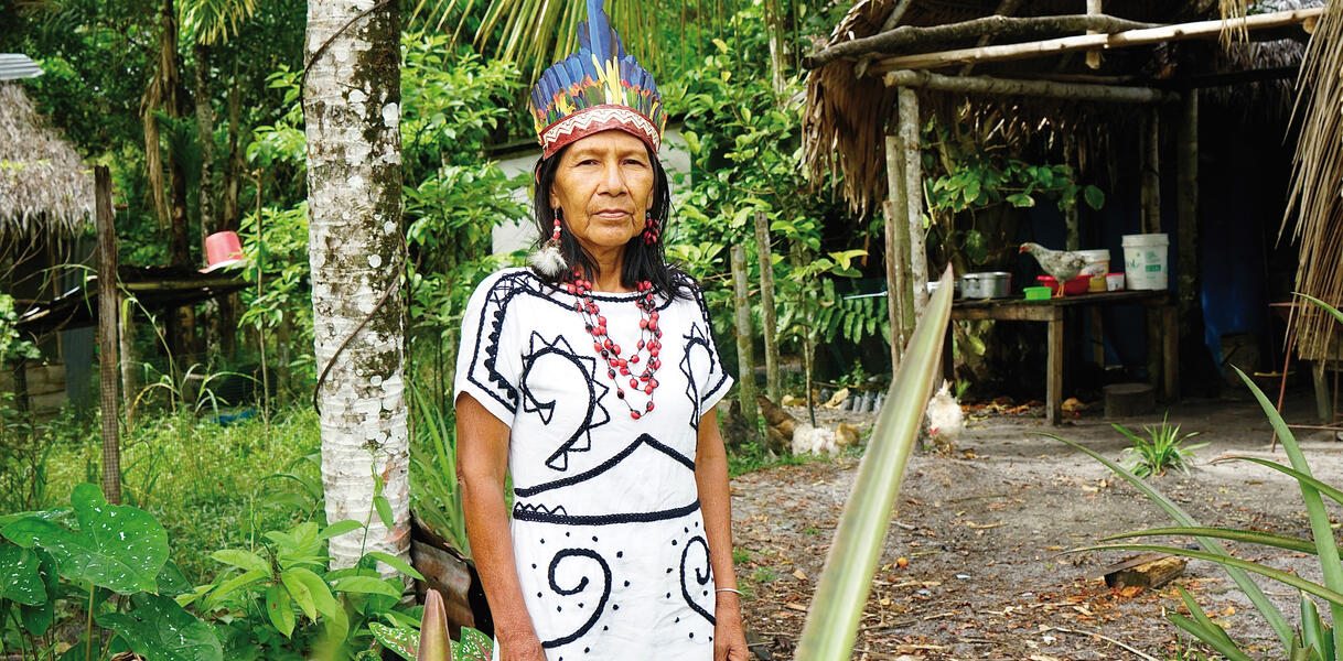 Eine indigene Frau mittleren Alters in tradtionellem Gewand, sie trägt eine Halskette und Federschmuck auf dem Kopf und steht im Wald vor einem Baum, im Hintergrund eine Hütte.