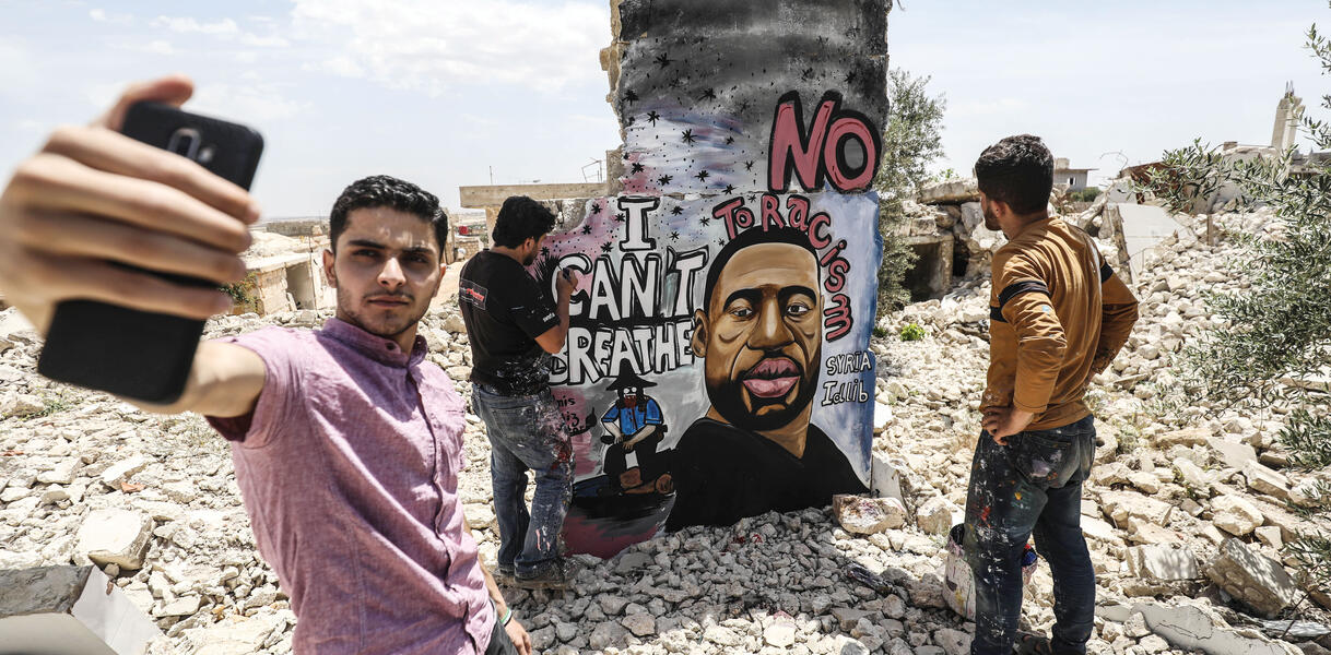 Junge Männer stehen inmitten der Ruine eines zerbombten Hauses, einer von ihnen malt ein Grafitti-Bild an einen Pfeiler, andere stehen um ihn herum, ein anderer macht ein Selfie mit seinem Handy.