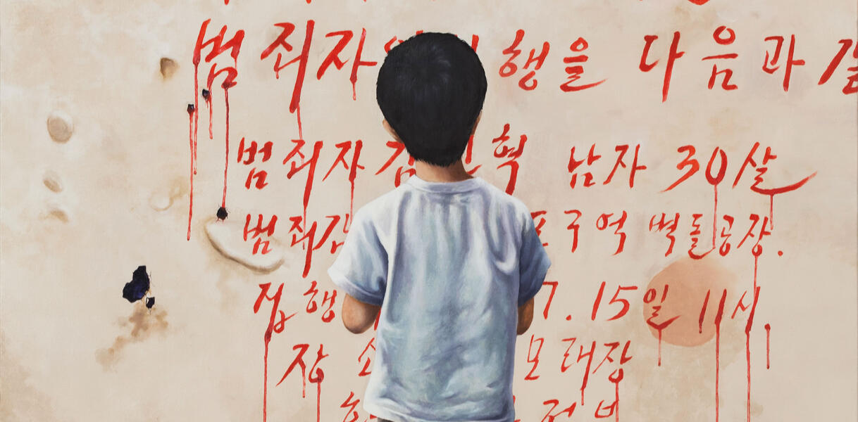Ein kleiner Junge steht vor einer Wand, auf die koreanische Schriftzeichen in roter Farbe gemalt sind, die nach unten hin verlaufen wie Blut. 