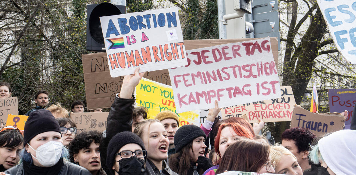 Viele junge Menschen stehen dicht gedrängt zusammen, manche haben einen Mund- und Nasenschutz auf. Eine Person hält ein Schild mit der Aufschrift "Abortion is a human right" hoch und ruft etwas. 