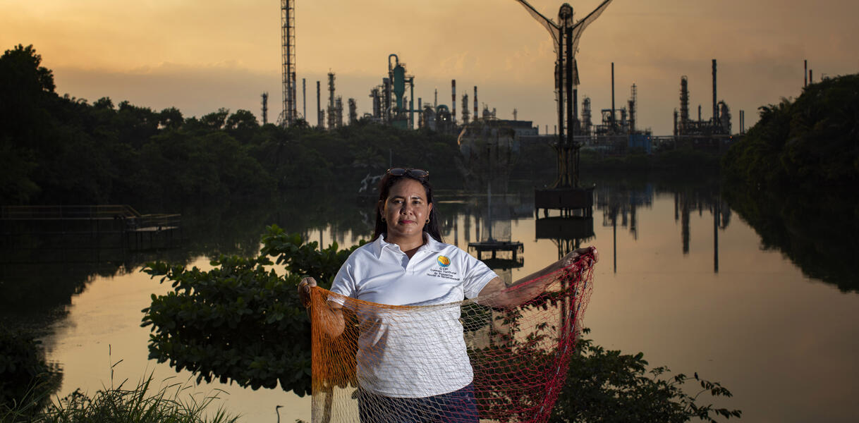 Das Bild zeigt eine Frau im Vordergrund, sie hält ein Fischernetz in der Hand, dahinter ist eine Industrieanlage zu sehen