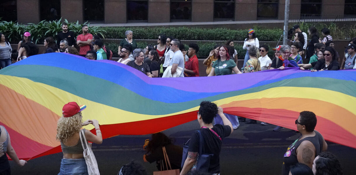 Das Bild zeigt Menschen, mit einen großen Regenbogenflagge