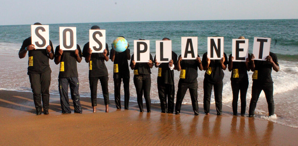 Das Foto zeigt zehn Personen, die jeweils Schilder mit Buchstaben vor ihre Gesichter halten. Dies ergibt den Schriftzug: "SOS Planet". Die vierte Person von rechts hält einen Globus vor ihr Gesicht.