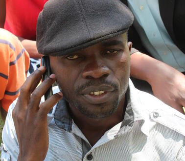 Das Foto zeigt Itai Peace Dzamara. Er trägt eine Mütze und hält ein Handy an sein rechtes Ohr. Hinter ihm stehen mehrere Personen.