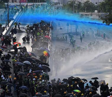 Demonstrierende Menschen, die sich mit Regenschirmen vor Angriffen der Polizei mit Wasserkanonen schützen. 