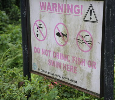 Weißes lädiertes Schild mit pinker Schrift und einem Ausrufezeichen in einem Dreieck, inmitten grüner Pflanzen. Text auf dem Schild: "Warning! Do not drink, fish or swim here"