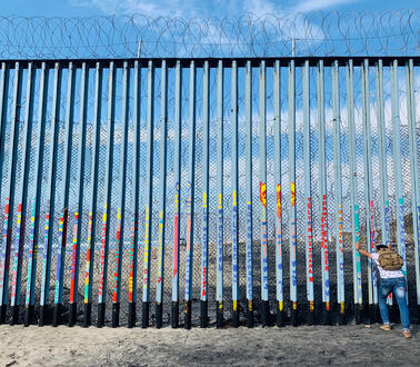 Ein meterhoher Grenzzaun aus Stahlstreben, darüber Stacheldrahtzaun, eine Person stemmt sich gegen den Zaun