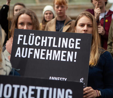 Zwei junge Frauen blicken in die Kamera und halten ein Schild in der Hand, auf dem steht: "Flüchtlinge aufnehmen". Hinter ihnen stehen weitere Personen, von denen einige ebenfalls Schilder hochhalten.