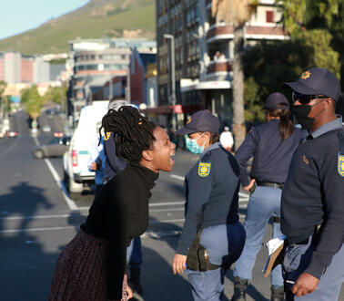 Eine Frau schreit auf einer Straße eine Gruppe Polizisten an, die Schirmmützen und Mund-Nase-Schutz tragen.