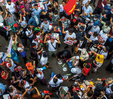 Aufnahme einer größeren Menschenmenge von oben. Einige der Demonstrierenden schwenken Fahnen und halten Plakate hoch. Auf einem der Plakate ist Auung San Suu Kyi abgebildet.
