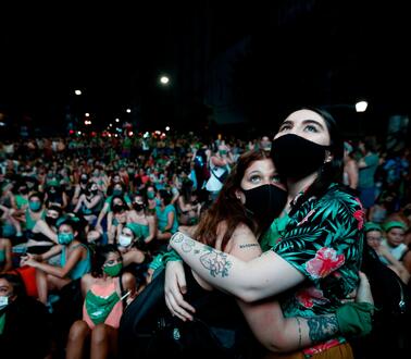 Zwei Frauen umarmen sich mit Masken im Vordergrund. Hinter ihnen befinden sich viele weitere Demonstrantinnen.