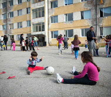 Kinder sitzen auf dem Boden und spielen Ball, im Hintergrund Menschen und ein Haus in schlechtem Zustand