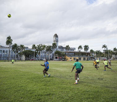 Menschen spielen Fußball auf einem Feld. Im Hintergrund stehen mehrere Gebäude, darunter eine Kirche.