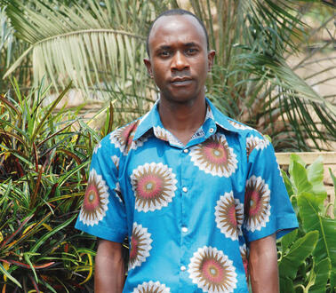 Ein junger Mann in einem kurzärmeligen Hemd steht vor einem Palmenhain und blickt in die Kamera.