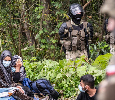 Zwei Frauen mit Kopftüchern und Mundschutz und ein Mann mit Mundschutz sitzen in einem Wald auf der Erde. Zwei uniformierte Soldaten mit Schutzhelmen stehen neben ihnen.