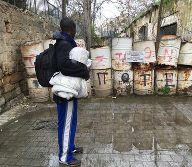 Ein Mann mit Rucksack und einer Decke unter dem Arm steht vor einem mit Tonnen und Stacheldraht versperrten Bereich auf einer Straße.