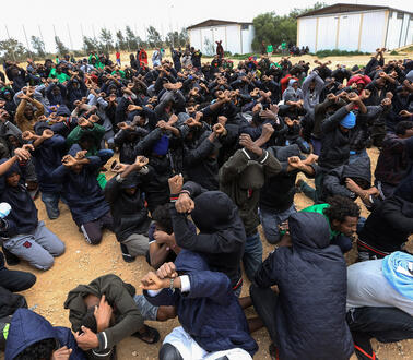 Das Bild zeigt viele Männer die mit gekreuzten Armen am Boden knien