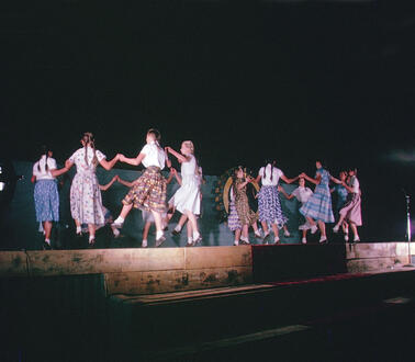 Mädchen in Blusen und Röcken halten sich an den Händen und tanzen einen Gruppentanz.
