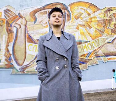 Das Bild zeigt eine junge Frau mit kurzen Haaren und Mantel vor einer Wand mit Grafitti
