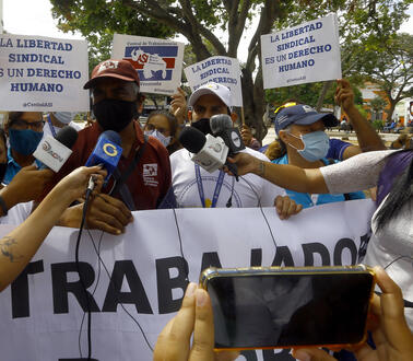 Das Bild zeigt die mehrere Menschen mit Schildern, davor stehen Reporter_innen mit Mikrofonen