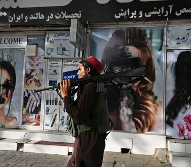 Ein Taliban-Kämpfer mit Sturmgewehr auf der Schulter geht an einem Geschäft vorbei. Auf den Plakaten des Geschäfts sind Frauengesichter schwarz übermalt.