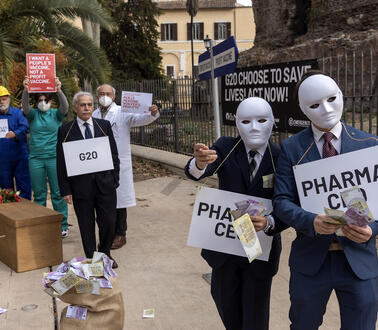 Sechs teilweise maskierte Personen halten auf einem Platz beschriftete Schilder in die Kamera.
