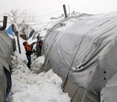 Ein Junge steht inmitten großer Schneemassen zwischen zwei Zelten und entfernt mit einem Besen Schnee von einem der Zelte. Zwei weitere Kinder stehen neben einem der Zelte und schauen ihm dabei zu.