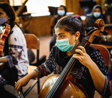 Eine junge Frau trägt einen Mundnasenschutz und spielt Cello, sie trägt traditionelle afghanische Kleidung, im Hintergrund sitzen weitere Frauen, die musizieren.