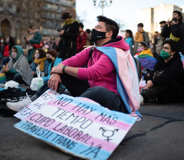 Menschen mit Schildern und Flaggen sitzen auf einem Platz. Im Vordergrund sitzt eine Person, die eine Transflagge umgebunden hat.